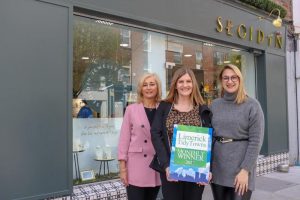 Three women standing outside shop. Middle holding award sign